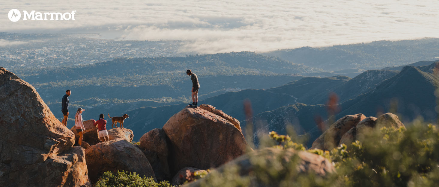 4 Marmot hikers and dog climb large rocks to look out at clouds and trees in background