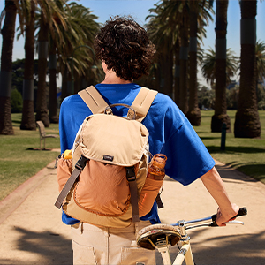 Two people wearing backpacks outdoors. One has a light blue backpack, the other an orange-brown backpack. Palm trees and structures visible in background.